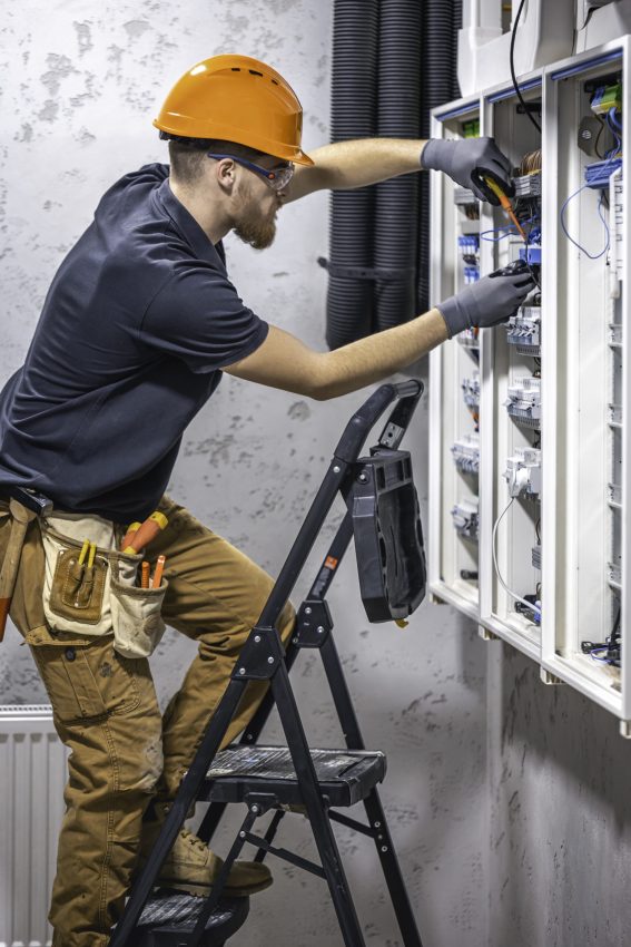 A male electrician works in a switchboard with an electrical connecting cable. Electrician with screwdriver tightens electrically operated switching equipment in fuse box.