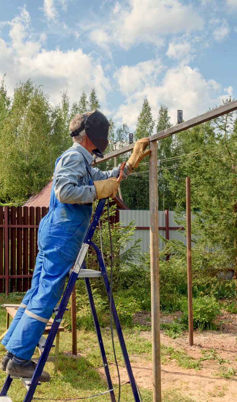 Man welder in a welding mask, construction uniform and protective gloves cooks metal on a street construction site. Construction of a pavilion, pergola near a country house on a summer day